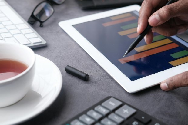 A close-up of a hand using a stylus on a tablet displaying bar graphs, surrounded by office items.