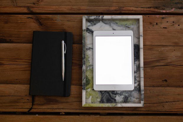Flat lay of a closed notebook with a pen next to a tablet on a marble tray on a wooden desk.